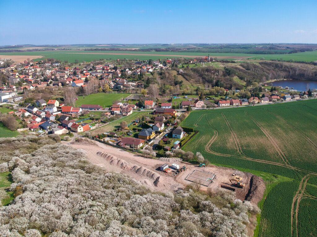 Vue aérienne en avril : le verger de mirabelliers en fleurs au premier plan, le terrain en cours de viabilisation, puis le village de Dřetovice et l'église Saint-Venceslas sur la colline